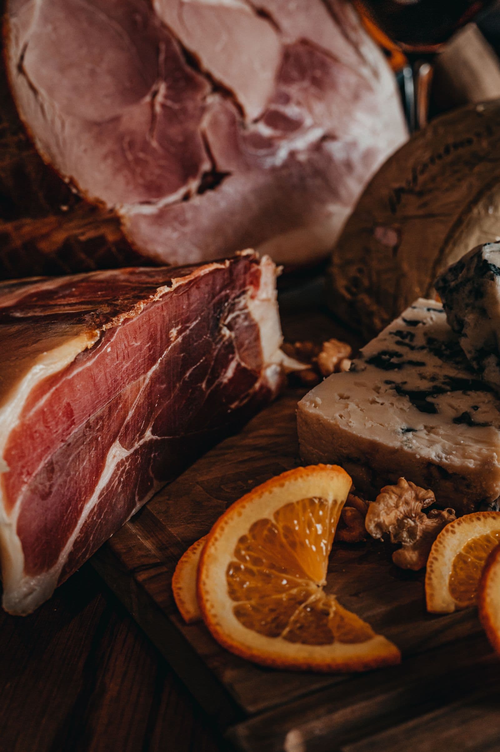 Un pezzo di prosciutto crudo al taglio su un tagliere di legno, accanto al gorgonzola, alle noci e a fette d'arancia essiccata